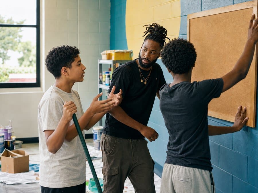 A mentor talking with two teenage boys while they hang a board on a wall